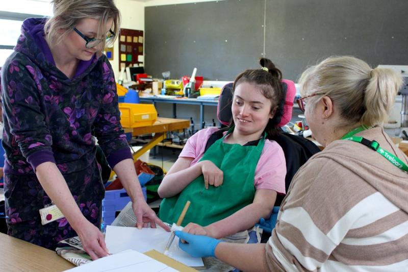 Treloar's student is using a paint brush to paint a stage prop; her assistant and teacher are standing next to her, assisting with the task: the assistant is holding her hand to help her paint and hold the brush. The teacher is holding the paper that's on the desk in front of the student.