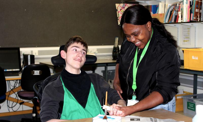 Treloar's student is using a paint brush to paint a stage prop; his assistant is holding his hand to help him paint and hold the brush. They are both smiling.