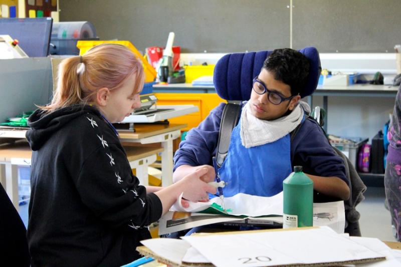 Treloar's student is using a paint brush to paint a stage prop; his assistant is holding his hand to help him paint and hold the brush. they are both really focused on the task.