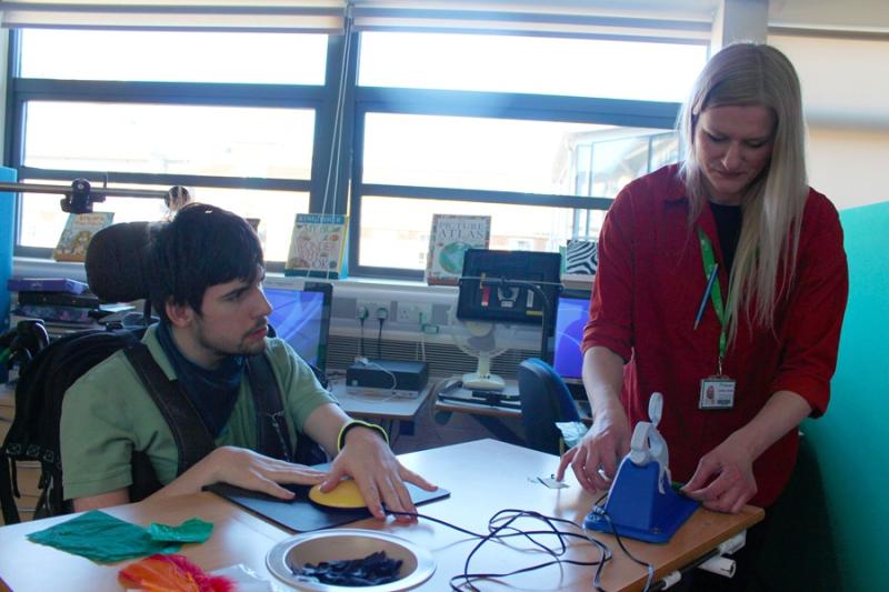 Treloar's student is using a yellow round switch to operate scissors which he is not able to access in a traditional way; his assistant is holding the piece of paper the student wants to cut.