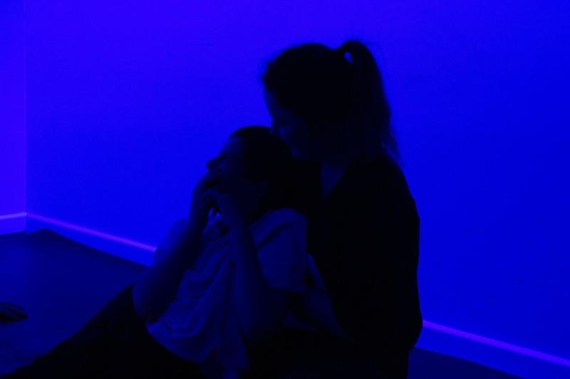 Treloar's student being held by his assistant - both sitting on a mat on the floor during an atmospherics session. The room is lit by a blue light. The student looks mesmerised by this sensory experience.