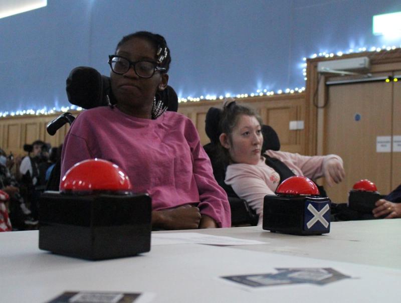 2 Treloar College students as judges during Treloar's Got Talent competition. They are watching the performance and on the desk in front of them are red buttons that they could press if they did not like the performance. In the background, there's audience watching the performers.
