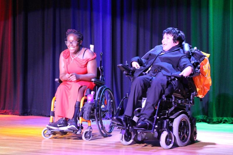 2 Treloar College students on the stage during Treloar's Got Talent competition. The stage is lit up with colourful lights. The students have just finished their performance and are looking at the audience, smiling. They are both smartly dressed: one is in a red dress, the other in a black suit.