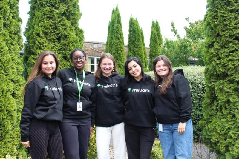 Five female volunteers standing outdoors in front of tall green shrubs, all wearing black hoodies with the 'Treloar's' logo on them.