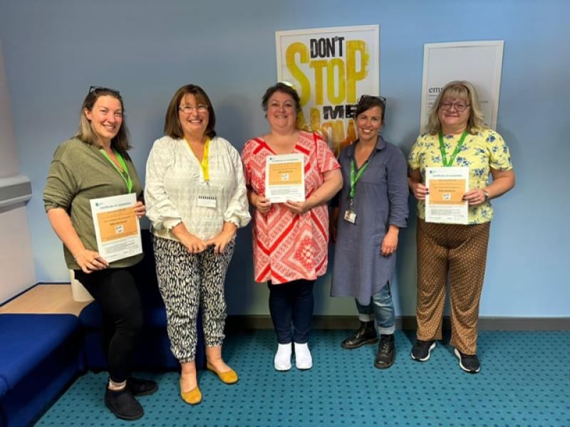 A group of 5 women, lined up in a room, posing for a picture. Some of them are holding certificates. Everyone is smiling.