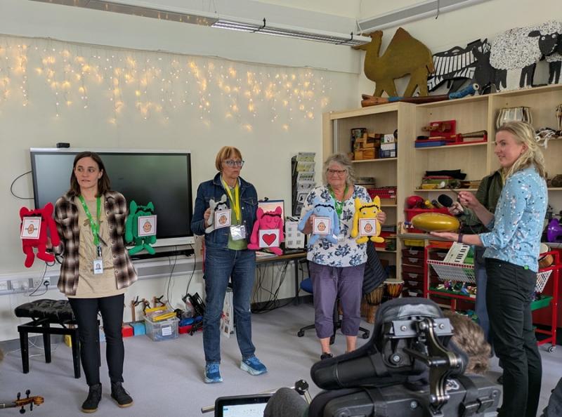 Philippa Perry from BBC Radio 3 with Treloar's staff members during a music lesson: they are all standing in front of the class, holding toys with attached stickers portraying different emotions.