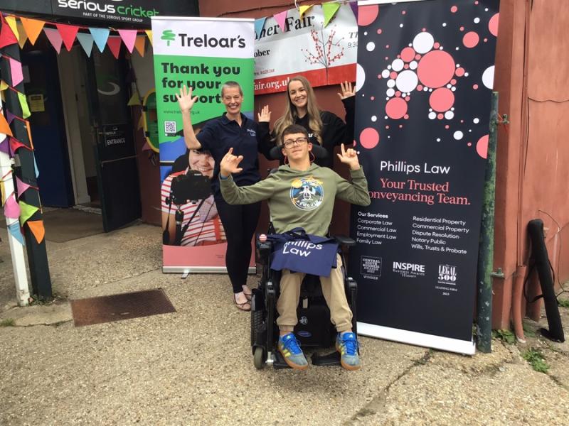 Treloar's student at the Dummer Fair posing for a photo with a sponsor and Treloar's member of staff in front of 2 banners. Everyone is waving at the camera.