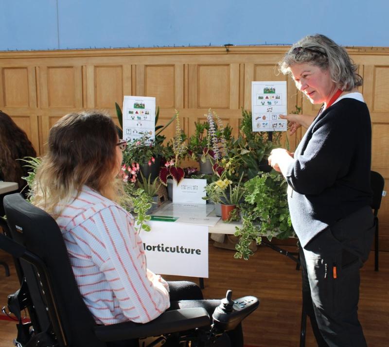 Treloar College student listens attentively to a member of staff explaining horticultural job opportunities; they are standing next to a table with various potted plants.