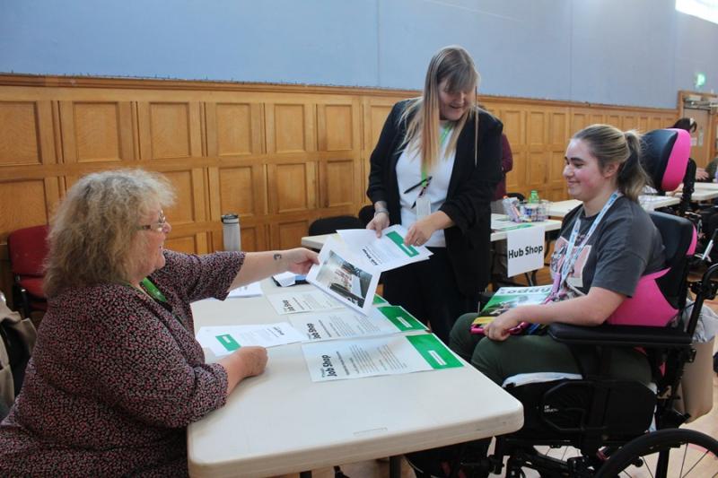 2 Treloar's staff members engage in a discussion with Treloar's student at a table covered with informational brochures inside a wood-paneled room.