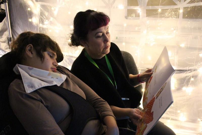 Treloar College student is being read a book by her assistant in a sensory tent with fairy lights on. The student is calm and looking at the illustrations in the book.
