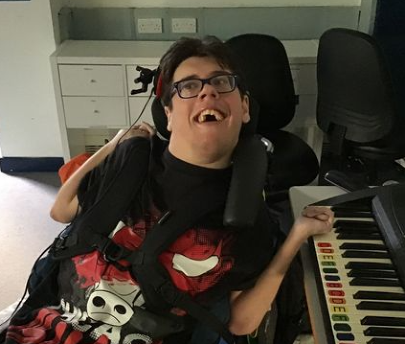 Treloar College student playing the piano with his left hand on the keys; he is wearing glasses and smiling at the camera.