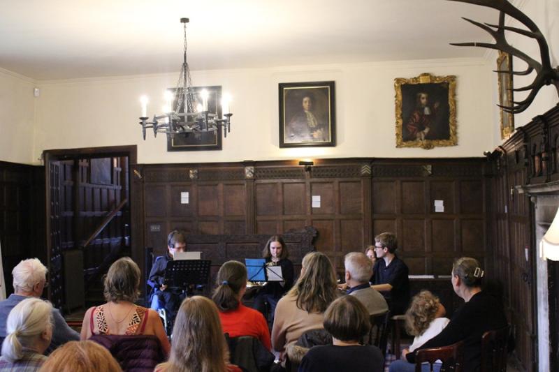 Treloar's former student Alessandro Vazzana performed with the Clarion Trio—Oscar Abbott on piano, Georgina Spray on French horn, and Alessandro on Clarion in a room in the Chawton House. An audience is sitting in front of the musicians and listening.