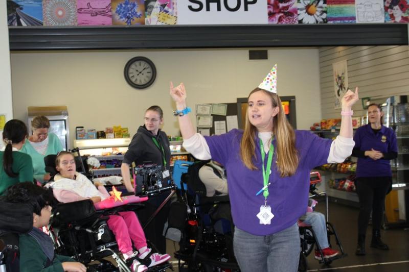 Treloar's Makaton tutor wearing a party hat and purple shirt joyfully raising her arms during a Makaton celebration event with with staff and students around her.