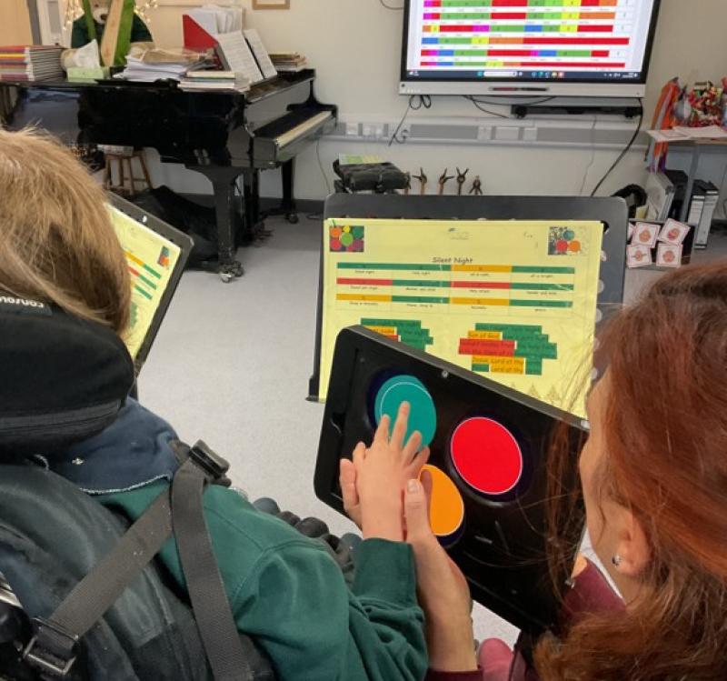 Over the shoulder shot of Treloar's student using a tablet with 3d printed case on which allows the student to play music using Clarion; the student is touching colored buttons on the screen to create music, she is guided by her assistant in a music classroom with a piano and sheet music on stands.