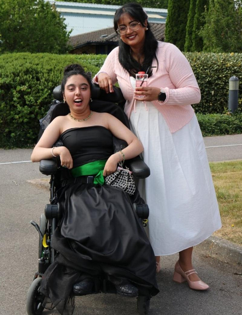 Treloar's student in a black strapless gown with a green belt outdoors during her prom, accompanied by her occupational therapist in a white dress and pink cardigan holding a drink.