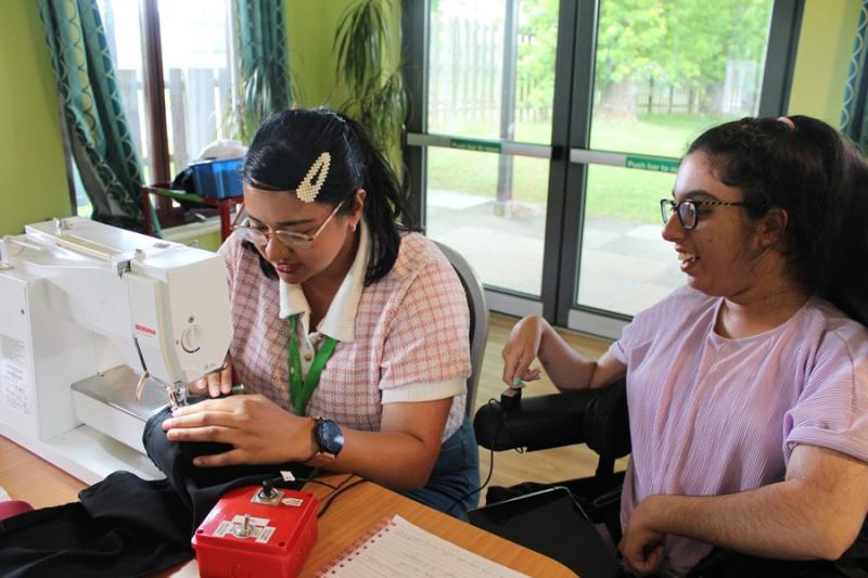 Treloar's occupational therapist operates a sewing machine, stitching fabric on a table in a bright room with large windows. Next to her is Treloar's student using a switch to power the sewing machine.