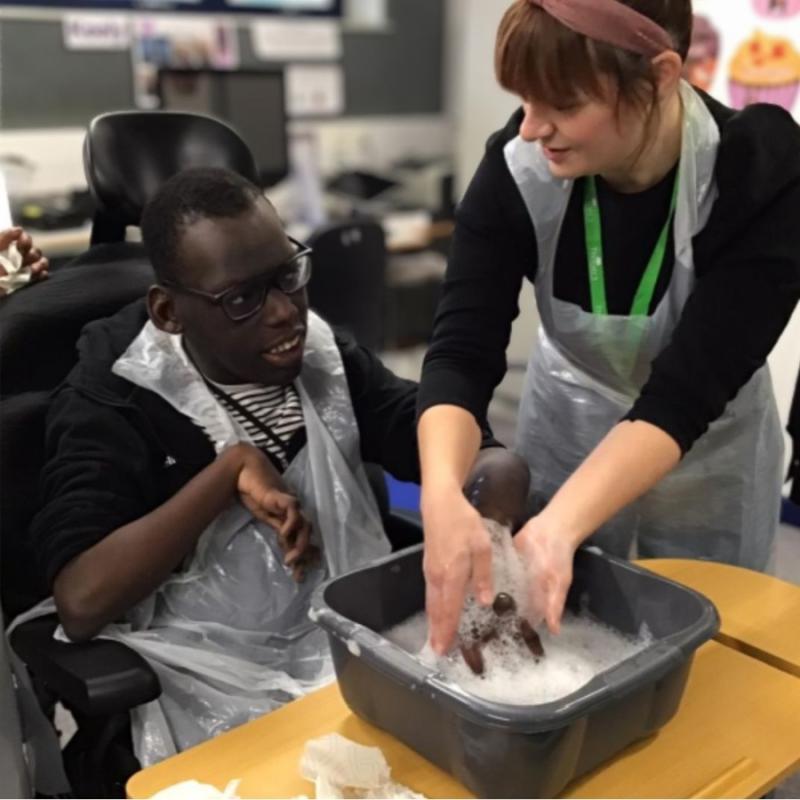 Treloar College student during his session with his occupational therapist: they are in the classroom and the OT is helping him wash his hand in a bowl with water with soap.