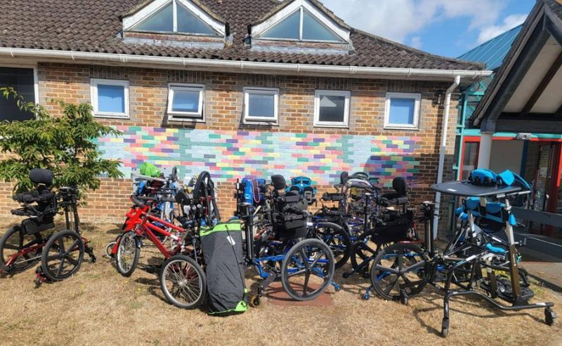A group of various adaptive bicycles and tricycles parked outside a brick building with colorful bricks on the wall.