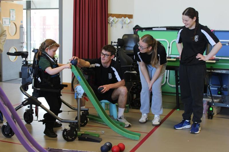 Treloar's primary student in the standing frame playing boccia with college students from a local college assisting her and observing her use a ramp.