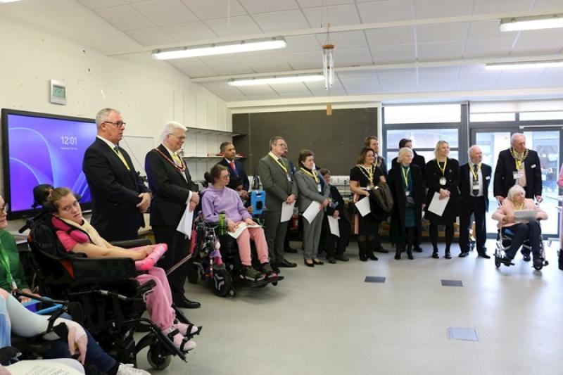 Group of people, including individuals in wheelchairs, standing and seated in a bright room during a formal event or ceremony.