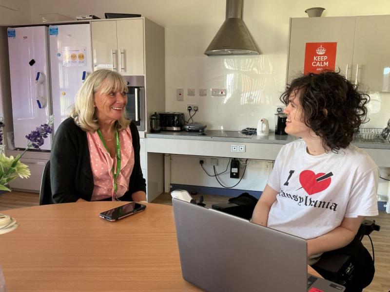 Treloar College student sits with a lap top round a table, in a kitchen interviewing a residential house manager.
