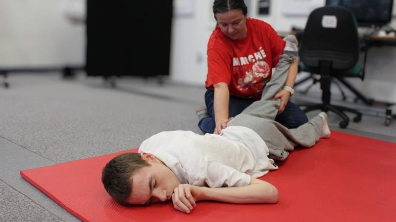 Treloar College student lying face down on a red mat while a staff member in a red shirt gently holds their leg, during a therapy / exercise session.