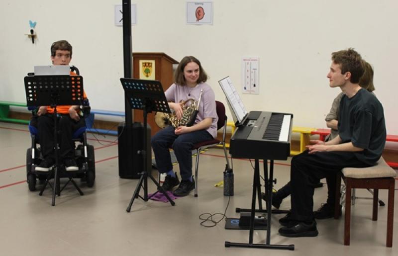 Three young musicians practicing indoors, one in a wheelchair with a music stand, another playing a French horn, and a third seated at an electronic keyboard.