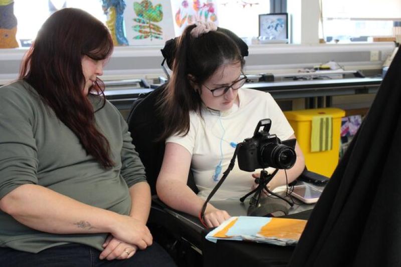 Treloar College student with her student support assistant seated in a classroom, with the student operating a camera on a tripod while the other watches attentively.