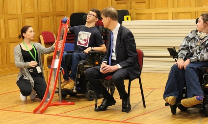 A support assistant kneels beside Treloar's student is demonstrating how to play boccia to Sir Stephen Timms who is wearing a suit and is watching attentively the student.