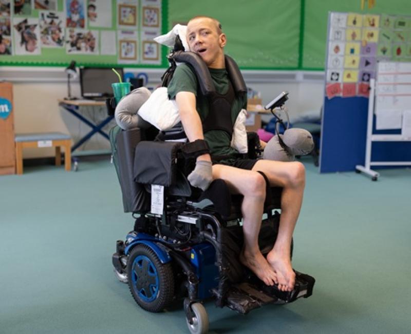 Treloar's student in his powered wheelchair which he drives using his foot; he is in a classroom setting with educational posters on the walls.