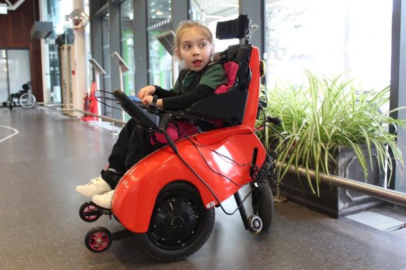 Treloar's primary student seated in a tiny trax chair wheelchair inside a spacious building with large windows and plants nearby.