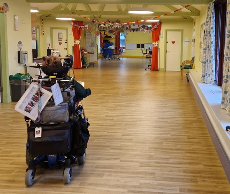 Treloar's student driving away in her wheelchair; she is moving down a wide, empty hallway with wooden floors and bright overhead lights.