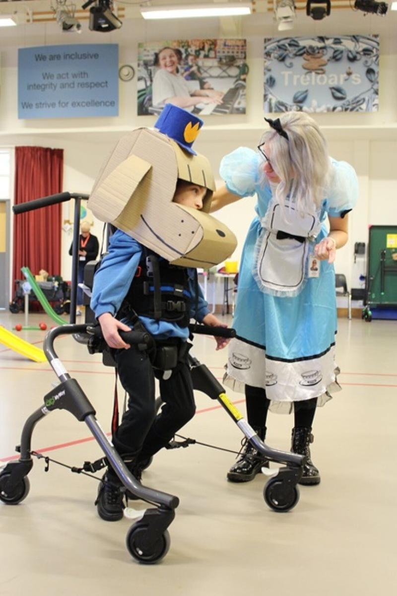 Treloar's primary student using a walker, wearing a large cardboard dog head and a hat, interacting with his Student Support Assistant dressed as Alice from Alice in Wonderland in a gymnasium.