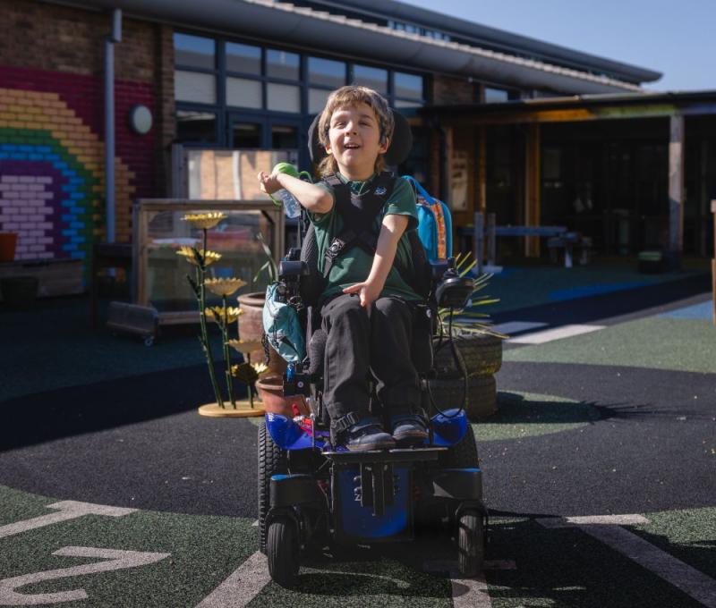 School student in the playground, in the sunshine in a power wheelchair with coloured bricks in a rainbow pattern behind them