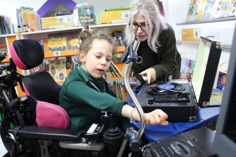 Treloar's primary student interacting with a record player while her assistant guides her in a colourful library setting.