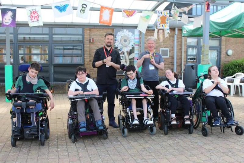 Group of 7 individuals, 5 Treloar's students and  2 adults outdoors under colourful hanging artwork in a courtyard.