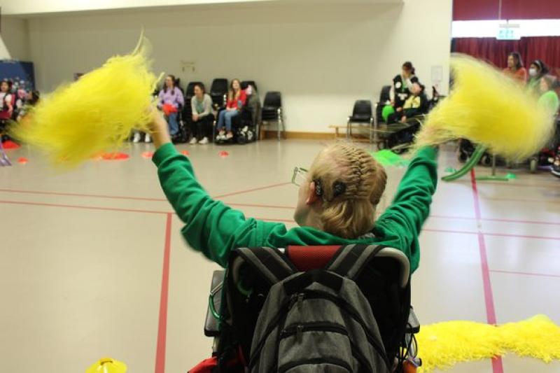 Treloar's student with braided hair enthusiastically waving yellow pom-poms in a gymnasium filled with seated spectators. He is wearing a cochlear implant.