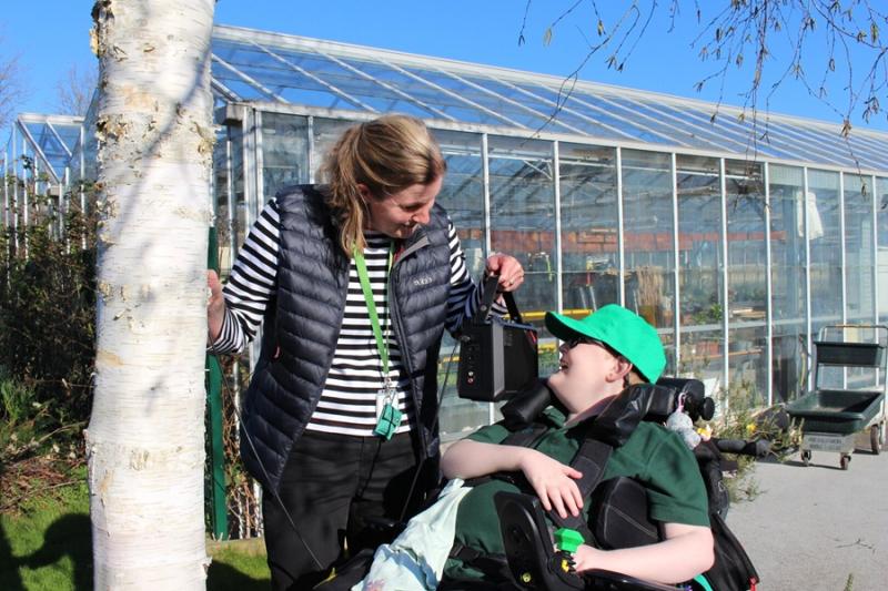Treloar's teacher in a striped shirt and black vest smiling and interacting with a student outside a large greenhouse on a sunny day. She is holding a speaker near the boys ear so that he can listen to the tree and the sounds of transpiration.