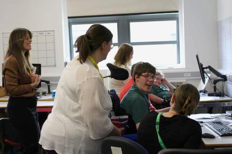 Two visitors with  yellow lanyards standing next to Treloar's students who are working on their computers in a classroom. One of the students is smiling and having a lively conversation with the visitor and his assistant.
