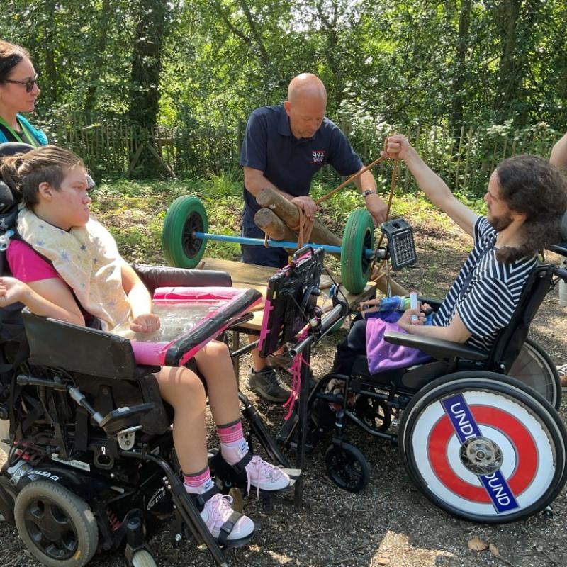 Two Treloar's students outdoors in the woods engaging with a man demonstrating a green-wheeled device in a wooded area. The students are looking carefully.