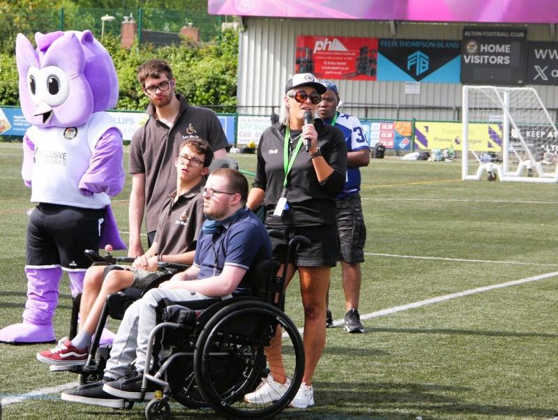 Group of people on a football field, including a Treloar's students and staff and a mascot in a purple costume, with one woman speaking into a microphone.