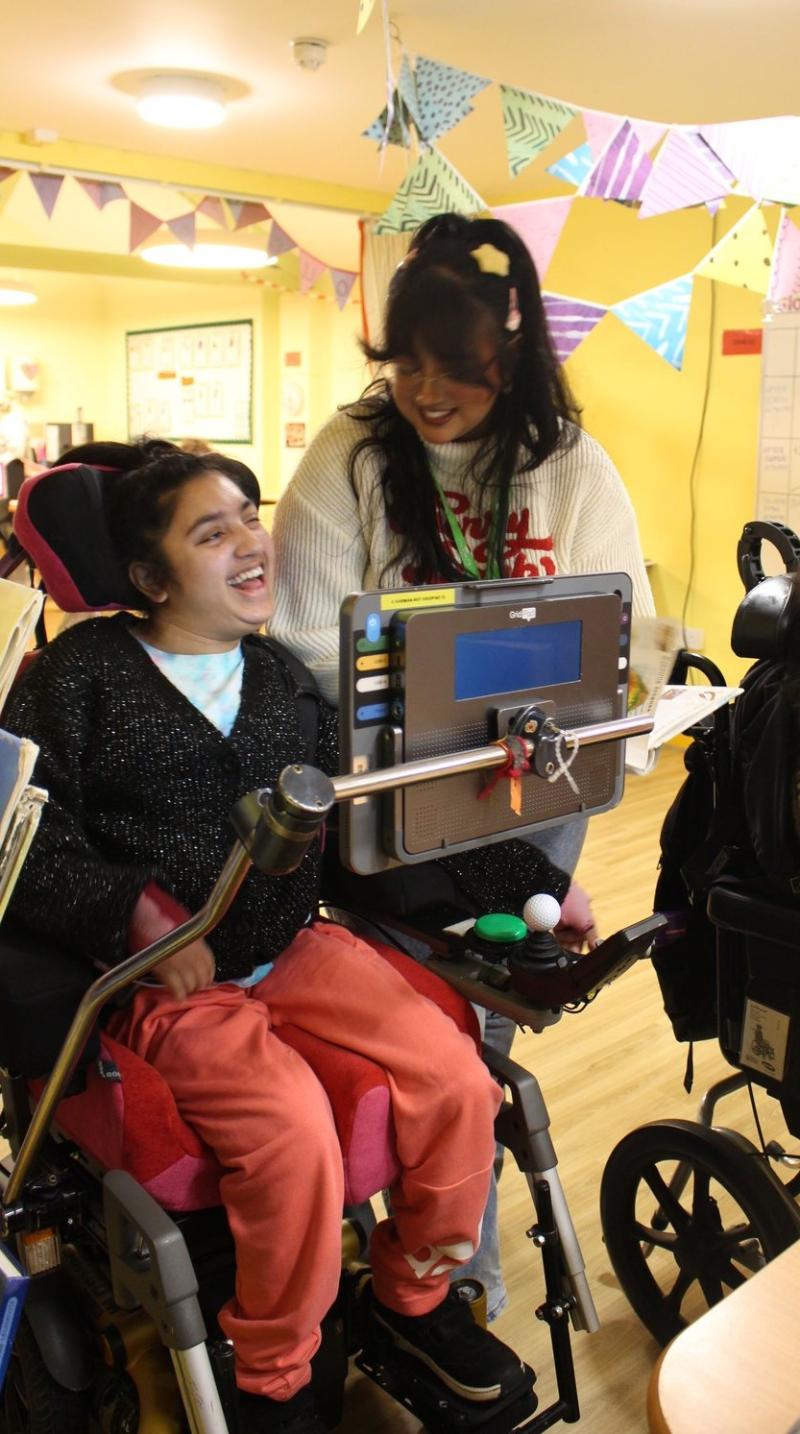 Treloar's student using a communication device in a decorated classroom with colourful bunting overhead. She is laughing together with her assistant who is standing right beside her.