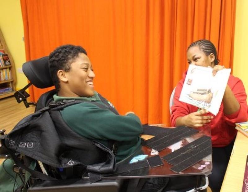 Treloar's student listens attentively to his assistant showing a book page in a room with orange curtains. He is smiling and enjoying the activity.