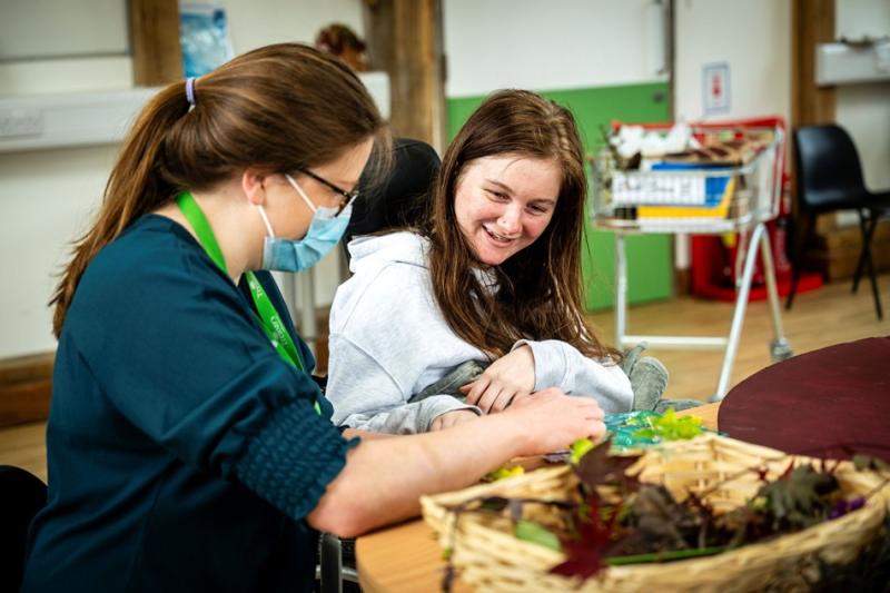 A student support assistant attentively engaging with Treloar's student during an indoor activity involving natural materials on a table. The student is happy to be taking part in the task.