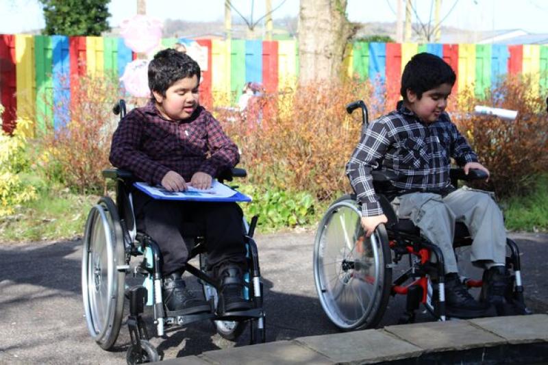 Two Treloar's students outdoors near a colourful fence, enjoying a sunny day in a garden setting. One of them has a clipboard with worksheets. Both focused on the pond in front of them.