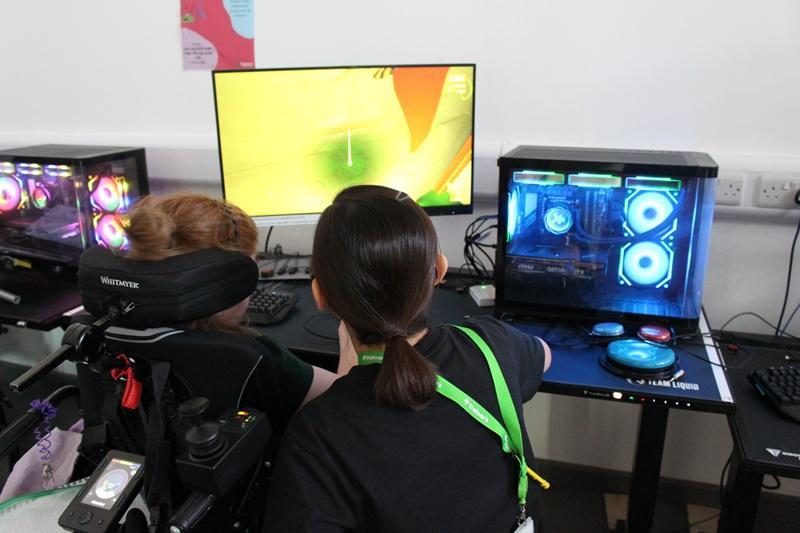 A support assistant assists Treloar's primary student playing a colourful video game on a desktop computer with illuminated cooling fans.