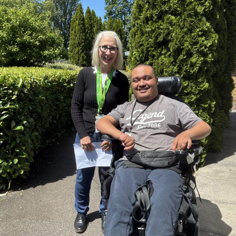 College tutor Flossie stands alongside Student in a wheelchair, both smiling, with trees and hedges behind them and clear blue skies