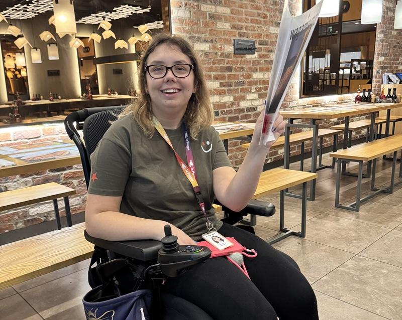 Treloar's student in an empty restaurant holding a big menu and smiling to the camera. She is wearing glasses and a work experience uniform.