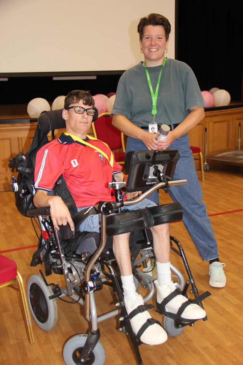 student in a wheel chair with a member of staff smiling towards the camera