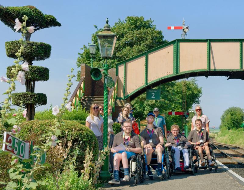Let Me In students visit The Watercress Line, a group of students in wheelchairs are in front of the railway bridge on the platform on a sunny day, the composition is framed with creative topiary